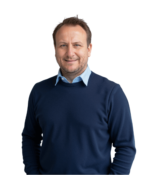 Man standing indoors in a professional setting, smiling at the camera while wearing a navy blue sweater over a light blue collared shirt.